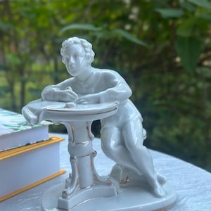 May include: A white porcelain figurine of a person sitting at a round table and writing. The figure has gold-painted trim on the table edges, the base, and the clothing details. The person is depicted in a seated position, leaning forward.