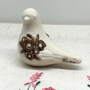 May include: A cream-coloured ceramic bird figurine with a brown floral design. The bird has a glossy finish and a tag that reads "Design Rosa Ljung". It rests on a surface with a floral pattern. A decorative item.