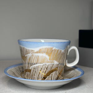 Puede incluir: Un juego de taza de té y platillo de porcelana blanca con un borde azul claro y un patrón metálico dorado repetitivo de tallos de trigo. La taza tiene un asa curva y el platillo muestra un diseño de trigo dorado a juego en su superficie.