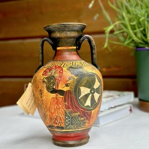 May include: A terracotta-colored ceramic amphora vase featuring two handles and a narrow neck. The body is decorated with black and red painted figures of a warrior holding a shield and helmet, accented by a Greek key pattern around the shoulder.