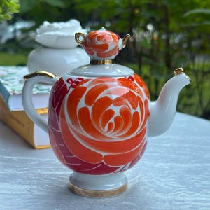 May include: A white porcelain teapot featuring a large, hand-painted orange and red rose motif. The teapot has a gold-trimmed handle, spout, and base, with a matching rose-shaped lid finial. The design includes bold, sweeping brushstrokes.