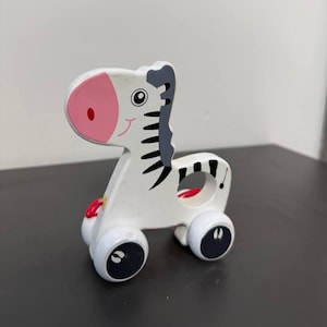 Op de afbeelding: Een wit houten speelgoedzebra op wielen met zwarte strepen, grijze manen en een roze snuit. De zebra heeft een cirkelvormige uitsparing in het midden van het lichaam en vier witte wielen met zwarte cirkelvormige details aan de zijkanten.