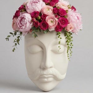 Peut inclure: Une jardinière murale en céramique blanc mat en forme de visage humain avec une moustache. La jardinière est remplie d'un arrangement dense de pivoines rose pâle, de roses magenta vibrantes, de petites renoncules et de feuilles d'eucalyptus.