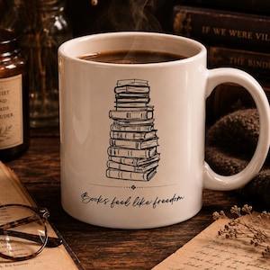Può includere: Una tazza in ceramica bianca riempita di liquido scuro fumante, caratterizzata da un disegno a linee nere di una pila verticale di libri e dal testo stampato "Books feel like freedom" in corsivo sotto l'illustrazione.