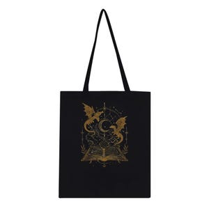 A flat black tote bag with two long shoulder straps features a gold foil illustration centered on the front showing an open book with two dragons and celestial symbols.