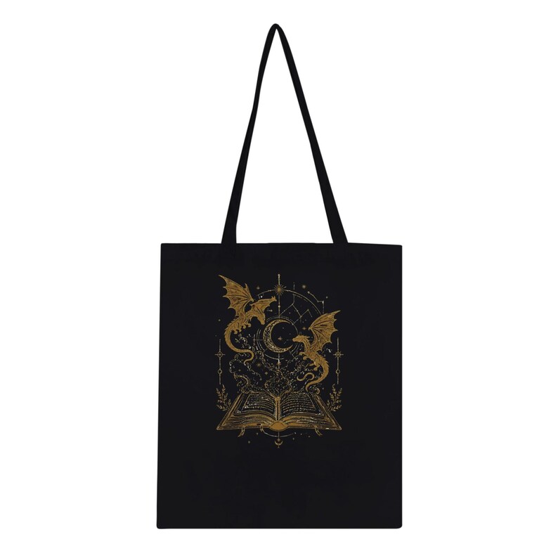 A flat black tote bag with two long shoulder straps features a gold foil illustration centered on the front showing an open book with two dragons and celestial symbols.