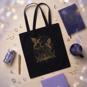 A black tote bag with a gold celestial dragon graphic lies on a light wooden background, encircled by purple notebooks, a cup of tea, and crystal beads.