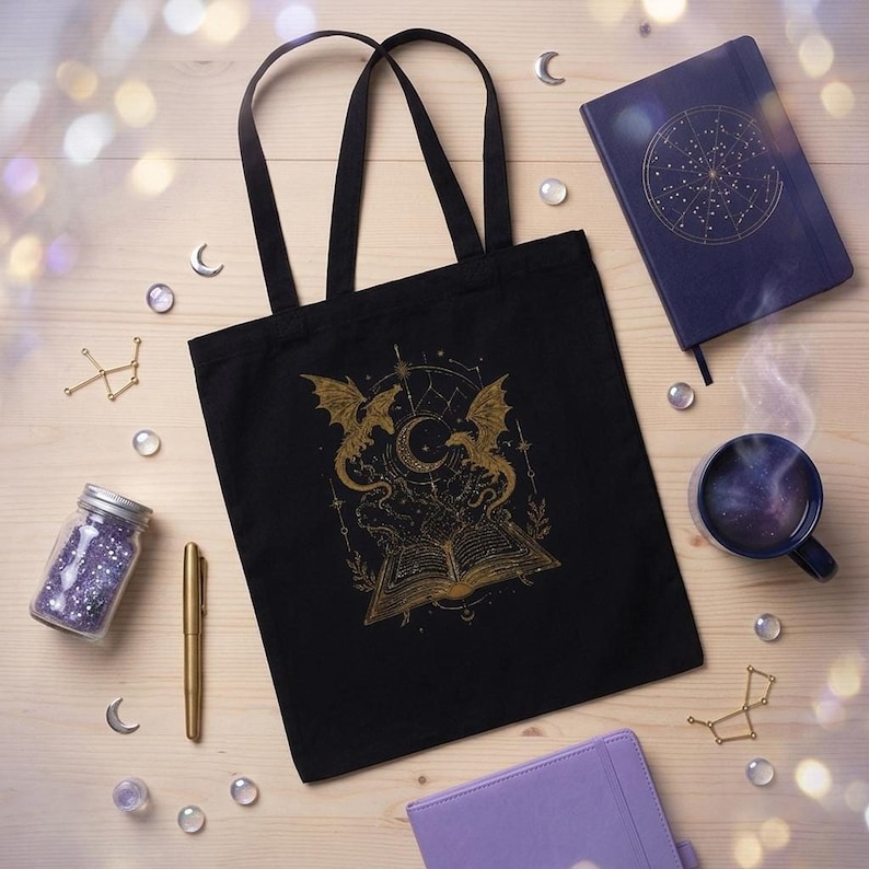 A black tote bag with a gold celestial dragon graphic lies on a light wooden background, encircled by purple notebooks, a cup of tea, and crystal beads.