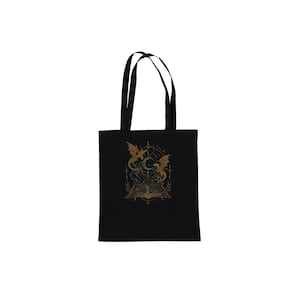 A black fabric tote bag with short handles displays a central gold graphic of a magical book and dragons against a plain white background.