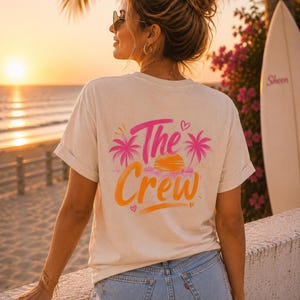 Könnte beinhalten: Ein cremefarbenes Kurzarm-T-Shirt mit einem Grafikdruck auf der Rückseite, der den Schriftzug The Crew in orangefarbener Schreibschrift zeigt, flankiert von zwei pinken Palmen und einer gelben Sonne sowie kleinen pinken Herzakzenten.