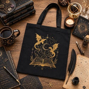 A black canvas tote bag lies flat on a dark wooden table, featuring a gold print of an open book, dragons, and a moon surrounded by celestial elements, with books and a mug nearby.
