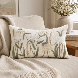 Può includere: Un cuscino rettangolare con una base bianco sporco caratterizzato da disegni botanici ricamati. Il ricamo include fiori bianchi, azzurri e beige con steli e foglie verde salvia, creando un motivo floreale dettagliato sulla parte anteriore.