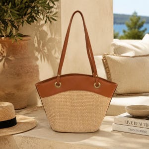 May include: A structured tote bag featuring a woven straw body in a natural beige tone and a smooth tan leather upper panel. The bag includes two long tan leather shoulder straps attached with gold-toned metal rings and hardware accents.