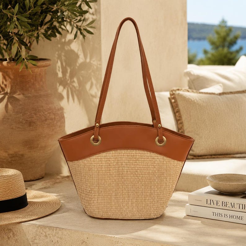May include: A structured tote bag featuring a woven straw body in a natural beige tone and a smooth tan leather upper panel. The bag includes two long tan leather shoulder straps attached with gold-toned metal rings and hardware accents.