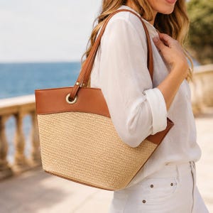 May include: A tote bag featuring a woven straw body in a natural beige tone, contrasted by smooth brown leather trim along the top edge and handles. The bag includes gold-toned metal grommets where the handles attach to the main structure.