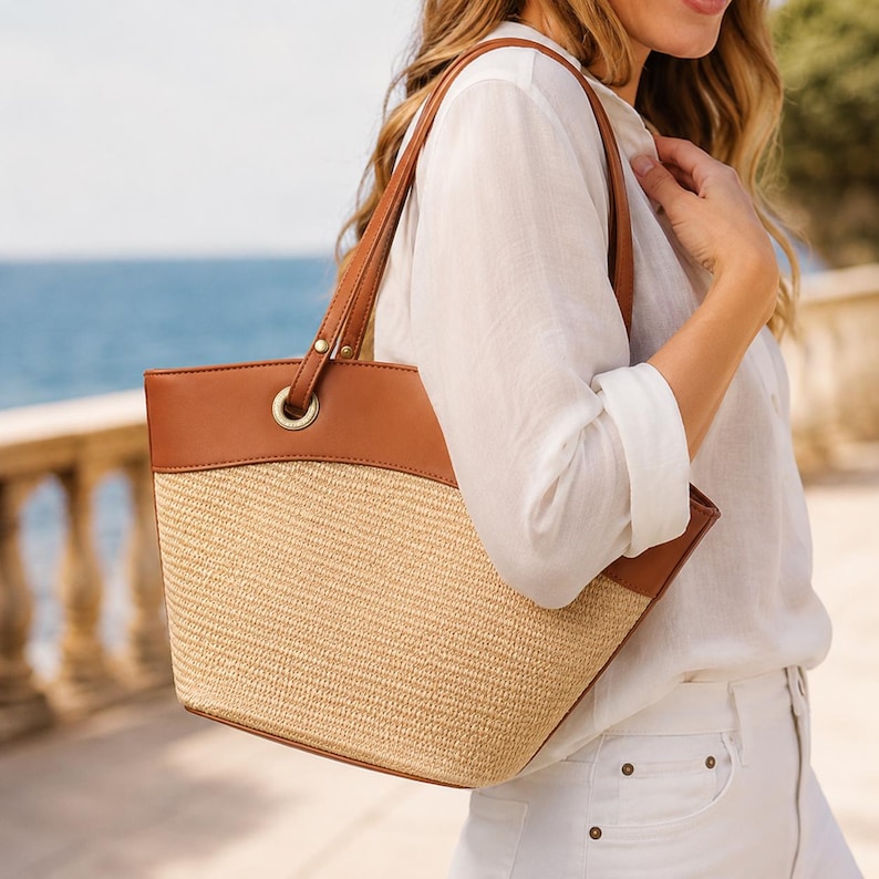 May include: A tote bag featuring a woven straw body in a natural beige tone, contrasted by smooth brown leather trim along the top edge and handles. The bag includes gold-toned metal grommets where the handles attach to the main structure.