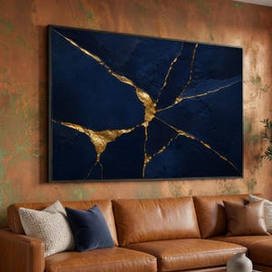 Op de afbeelding: Groot ingelijst abstract kunstwerk. Het kunstwerk heeft een diep donkerblauwe achtergrond met goudkleurige scheurachtige lijnen. Het stuk is rechthoekig en gemonteerd in een zwart frame. De kunst wordt boven een bruine leren bank getoond.