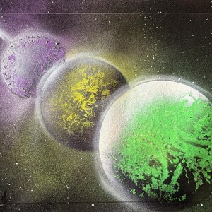 May include: An abstract painting of three planets in space. The first planet is purple, the second is yellow, and the third is green. White streaks and stars are scattered throughout the dark background, creating a cosmic effect.