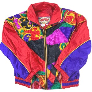 May include: A zip-up jacket featuring a vibrant, multi-colored patchwork design in red, purple, black, green, and gold. The garment includes a high collar, long sleeves with elastic cuffs, and a label inside the neck that reads East West.