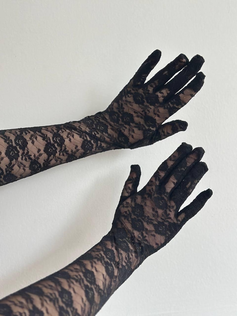 May include: Black lace gloves with a floral pattern. The gloves are long, extending up the arm, and are made of a delicate, see-through fabric. The background is a plain white wall.