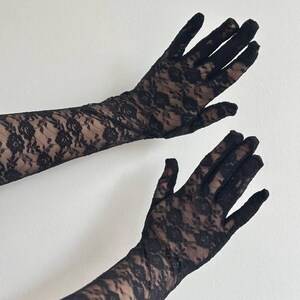 May include: Black lace gloves with a floral pattern. The gloves are long, extending past the wrists, and are made of a delicate, see-through fabric. The background is a plain white wall.