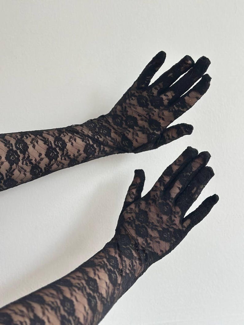 May include: Black lace gloves with a floral pattern. The gloves are long, extending past the wrists, and are made of a delicate, see-through fabric. The background is a plain white wall.