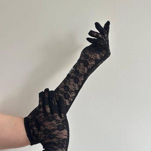 May include: Black lace gloves with a floral pattern. The gloves are long, extending past the wrist, and are made of a delicate, see-through fabric. The gloves are shown against a plain, off-white background.
