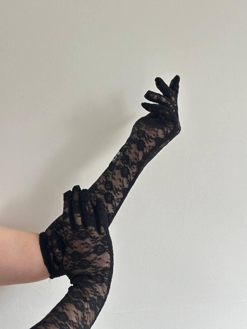 May include: Black lace gloves with a floral pattern. The gloves are long, extending past the wrist, and are made of a delicate, see-through fabric. The gloves are shown against a plain, off-white background.