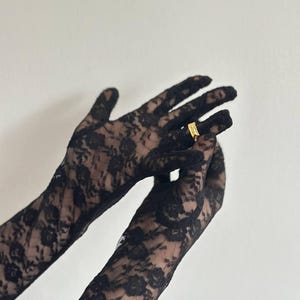 May include: Black lace gloves with a floral pattern. A gold ring is on one finger. The gloves are long and cover the hands and wrists.