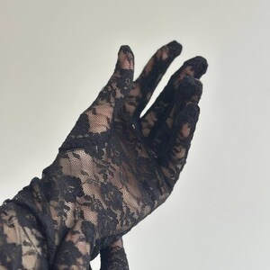 May include: Black lace gloves with a floral pattern. The gloves are made of a delicate, sheer fabric, and the intricate design is visible against a light background. The gloves extend up the arm.