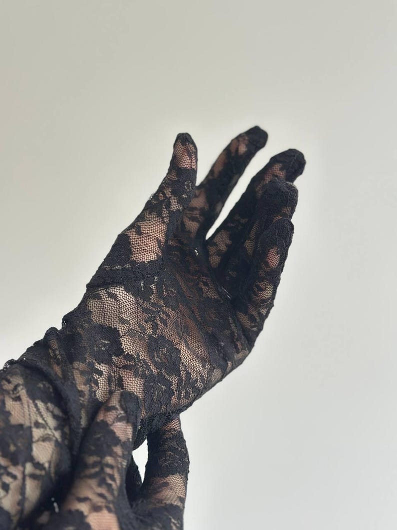 May include: Black lace gloves with a floral pattern. The gloves are made of a delicate, sheer fabric, and the intricate design is visible against a light background. The gloves extend up the arm.