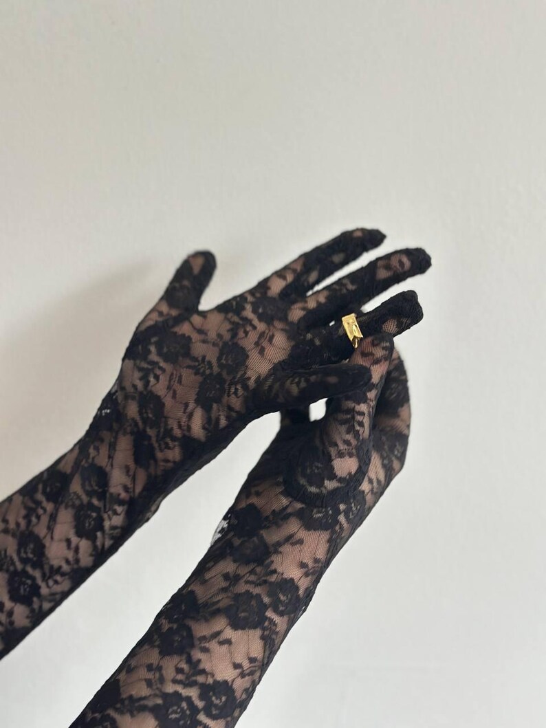 May include: Black lace gloves with a floral pattern. A gold ring is on one finger. The gloves are long and cover the hands and wrists.
