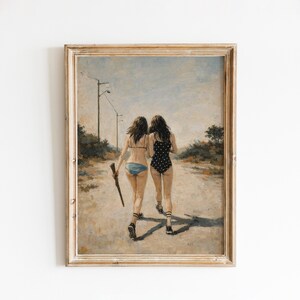 Op de afbeelding: Ingelijste kunstdruk met twee figuren die weglopen op een onverharde weg. Eén figuur draagt een blauwe bikini, de ander een zwart-wit polkadot badpak. Eén figuur draagt een lang, donker object. Het kunstwerk is in vintage stijl.