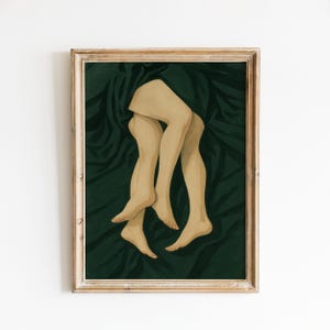 May include: A framed art print featuring a stylized illustration of two pairs of legs intertwined against a dark forest green background with deep, flowing fabric folds. The artwork is encased in a light-toned wooden frame with a visible grain.