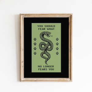 Puede incluir: Una lámina de arte enmarcada con un fondo rectangular verde salvia y una ilustración de serpiente pixelada negra en el centro. El texto dice YOU SHOULD FEAR WHAT sobre la serpiente y NO LONGER FEARS YOU debajo, con pequeños iconos florales.