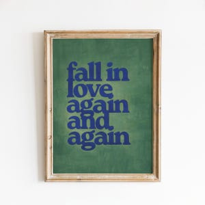May include: A framed print featuring the phrase "fall in love again and again" in blue text against a green backdrop. The frame is a light brown colour.
