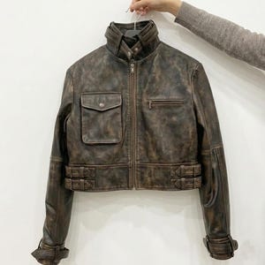 May include: A cropped brown leather jacket with a distressed finish, featuring a high collar with a snap closure, a front zipper, one flap pocket on the right chest, one zippered pocket on the left chest, and adjustable buckle straps at the waist.