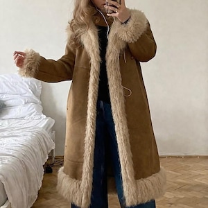 May include: A long, tan suede-like coat featuring a thick, cream-colored faux fur trim along the wide lapels, cuffs, and bottom hem. The coat is worn over a black top and dark blue wide-leg denim jeans, creating a layered look with visible texture.