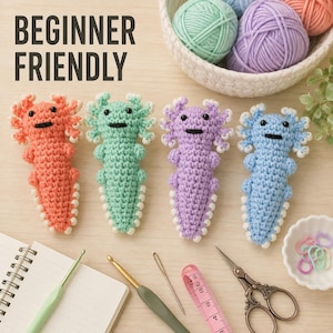 Könnte beinhalten: Vier gehäkelte Axolotl-Stofftiere in Koralle, Mintgrün, Lavendel und Hellblau mit schwarzen Augen und weißen Kiemen. Der Text BEGINNER FRIENDLY ist zu sehen. Bastelzubehör umfasst Notizbuch, Häkelnadeln, Nadel, Schere und ein Maßband.