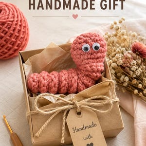 May include: A pink crocheted worm toy with googly eyes inside a brown cardboard gift box. The box is tied with twine and features a tag that reads "Handmade with" followed by a heart icon. The text "ADORABLE HANDMADE GIFT" appears above.