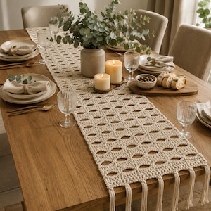 Puede incluir: Un camino de mesa de ganchillo color crema con un patrón ovalado calado y largos flecos en los extremos. El camino está sobre una mesa de comedor de madera con platos, servilletas, cristalería, un jarrón con eucalipto y dos velas.