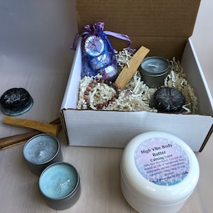 May include: A white box filled with a variety of items, including candles, a crystal, a bag of crystals, and a jar of body butter. The body butter jar has a label that reads "High Vibe Body Butter Calming Love 100% Raw Shea Butter, Coconut Oil,  Almond Oil,  Oil of Sandalwood, Frankincense, Clary Sage, Ylang Ylang, Cardamom, Jasmine, Vanilla, Orange Blossom,  Yang Yang Orange  Made with love for your body"