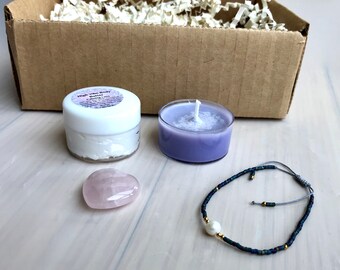 May include: A self-care gift box with a small jar of body butter labeled "High Vibe Body Butter Calming Love", a purple candle, a pink heart-shaped crystal, and a blue beaded bracelet with a pearl charm.