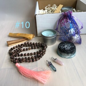 May include: A gift box filled with items for self-care and relaxation. The box includes a candle, a soap bar, a mala necklace with a pink tassel, two crystal pendants, two sticks of palo santo wood, and a small bag of white stones.