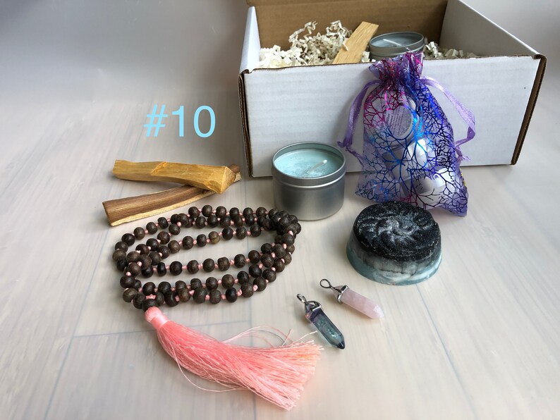 May include: A gift box filled with items for self-care and relaxation. The box includes a candle, a soap bar, a mala necklace with a pink tassel, two crystal pendants, two sticks of palo santo wood, and a small bag of white stones.