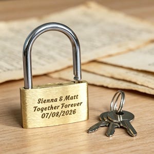 Peut inclure: Un cadenas en laiton avec une anse argentée, gravé avec le texte Sienna & Matt, Together Forever, et la date 07/08/2026. Le cadenas est ouvert et un trousseau de trois clés argentées sur un anneau métallique est posé à côté.