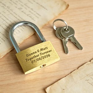 May include: A brass padlock with a silver-coloured shackle and a set of two keys on a ring. The padlock is engraved with the text: Sienna & Matt, Together Forever, 07/08/2026. The metal surface has a brushed finish and a visible keyhole at the bottom.