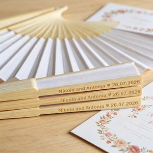 Peut inclure: Trois éventails pliés avec des montures en bois clair et du tissu blanc. Chaque manche est gravé avec le texte Nicola and Antonia, un symbole de cœur et la date 26.07.2026. Un éventail ouvert et de la papeterie florale sont visibles en arrière-plan.