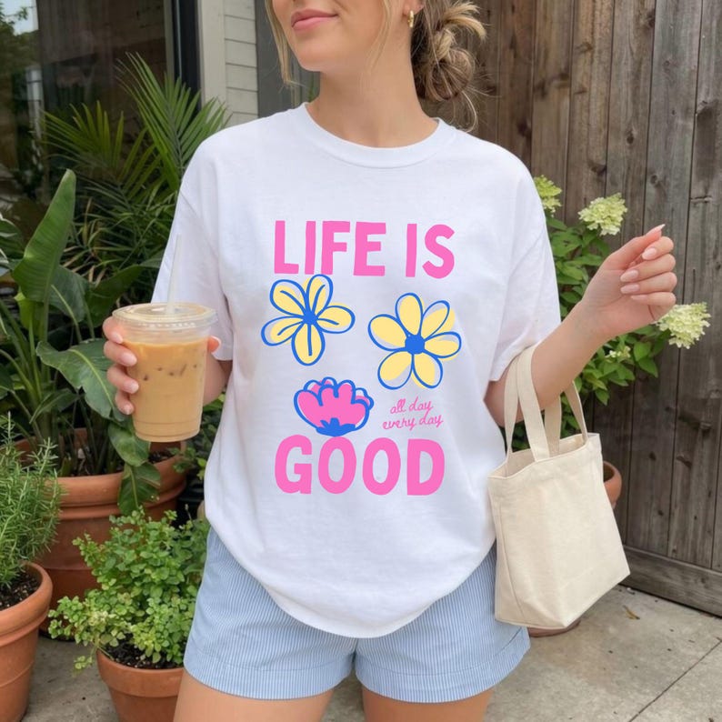 May include: White t-shirt with the text "LIFE IS GOOD" in pink, and flower graphics in blue and yellow. The shirt is worn with light blue shorts and a small canvas tote bag. A clear plastic cup with iced coffee is held in the left hand.