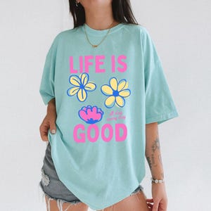May include: A pale blue t-shirt featuring the pink text "LIFE IS GOOD" and three flower illustrations. The shirt has a crew neck and short sleeves. The words "all day everyday" are also printed on the shirt.